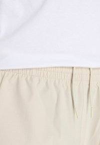 Pantalones cortos beige de cintura elástica con una textura suave y cordón ajustable, llevados bajo una camiseta blanca sencilla.
