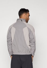 Pelēks vējjaka ar elastīgām piedurknēm un apakšmalu, ar kontrastējošiem gaiši pelēkiem paneļu akcentiem un augstu apkakli.