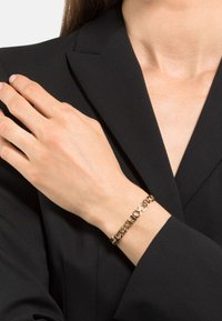 Pulsera en tono dorado con un diseño alternado de óvalos y eslabones. Lleva puesta en una muñeca, complementando un blazer oscuro. Acabado metálico suave.