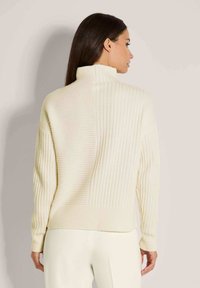 Vrouw met lang, donker haar die een off-white geribbelde trui met hoge hals draagt, gezien vanuit de rug tegen een effen achtergrond.