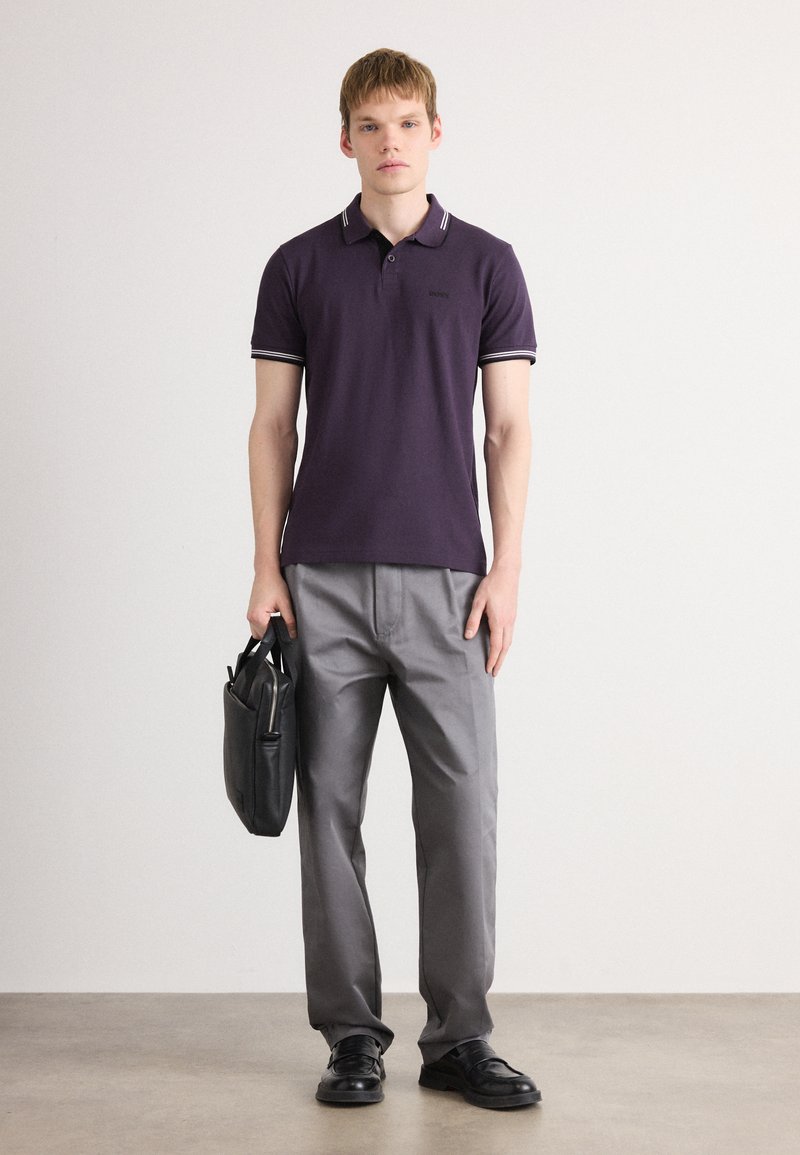 Jeune homme portant un polo violet foncé, un pantalon gris, des chaussures noires, tenant une mallette noire devant un mur blanc uni.