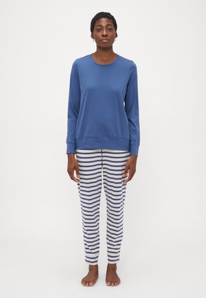 Donna in piedi a piedi nudi, con una camicia blu a maniche lunghe e pantaloni bianchi con strisce orizzontali blu, contro uno sfondo bianco semplice.