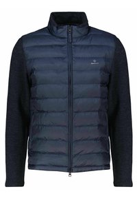 Navy padded vest with zip closure, featuring a textured dark gray knit fabric on sleeves and collar, and a small logo on the chest.