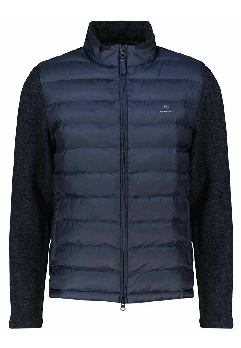 Navy padded vest with zip closure, featuring a textured dark gray knit fabric on sleeves and collar, and a small logo on the chest.