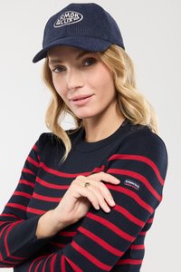 Granatowy kapelusz z welurową teksturą i białym logo. Sweterek dzianinowy w czerwono-granatowe paski z okrągłym dekoltem i małym detalem metki na rękawie.