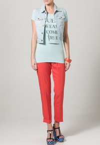 Jeansweste mit goldenen Knöpfen über einem hellblauen Grafik-T-Shirt. Helle korallfarbene Hose und blaue Plateausandalen mit Punkten vervollständigen das Outfit.