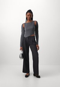 Grau gerippter Langarmoberteil mit Cut-Out-Details, kombiniert mit hoch-taillierten, schwarzen, weiten Hosen und schwarzen Slippers, während eine silberne Handtasche gehalten wird.