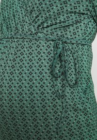 Zelené vzorované šaty z látky s černým geometrickým designem, na guma v pase a s tkaničkou. Měkká textura s mírným leskem.