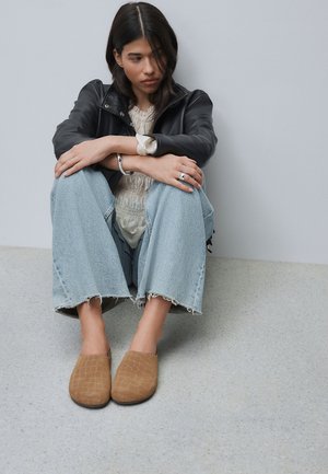 Frau sitzt auf dem Boden an der Wand gelehnt mit verschränkten Armen auf den Knien, trägt eine schwarze Lederjacke, hellblaue Jeans und braune Slipper.