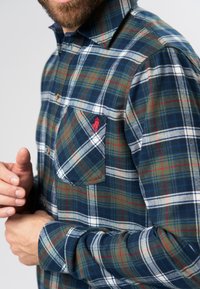Gevlekt flanellen shirt in blauw, groen en wit met rode accenten. Heeft een button-down kraag en een borstzak met een logo-embleem.