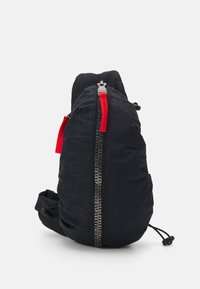 Sac à dos noir avec une surface texturée, présentant une fermeture éclair rouge et un détail de texte graphique. Design compact et épuré avec une sangle réglable.