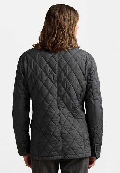 Person mit schulterlangem braunem Haar, der eine dunkelgraue gesteppte Jacke trägt, von hinten gegen einen weißen Hintergrund gezeigt.