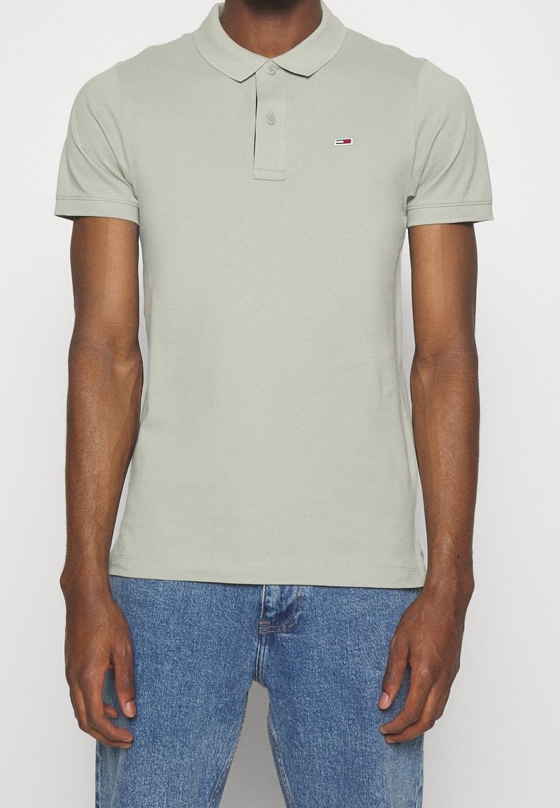 Homme portant un polo gris clair à manches courtes avec petit logo rouge et blanc sur la poitrine et un jean bleu.