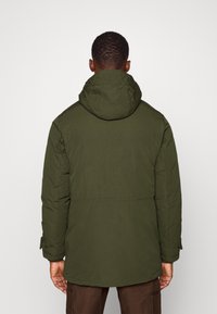 Mörkgrön vadderad jacka med huva, med en slät textur, justerbara ärmslut och en mittlång design. Sömmar synliga.