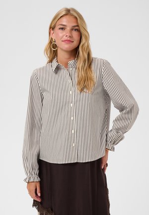 Femme aux longs cheveux blonds portant de grandes boucles d'oreilles en cercle, une chemise blanche à rayures verticales noires et une jupe foncée avec un bord en dentelle.
