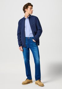 Marinblå zip-up jacka över en ljusblå skjorta med knappar, kombinerad med blå jeans och beige skor. Slät textur och avslappnad design.