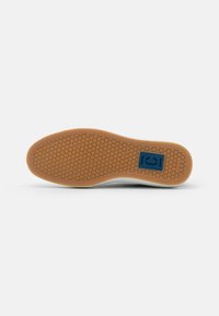 Gumová podrážka s krajkovým vzorem, na které je modré logo. Podrážka je světle hnědá s texturovaným povrchem pro lepší přilnavost.