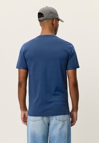 Μπλε μπλουζάκι από βαμβάκι με κοντά μανίκια, στρογγυλή λαιμόκοψη και καθαρό φινίρισμα. Φοριέται με ανοιχτό μπλε τζιν και γκρι καπέλο.