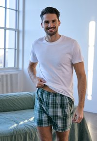 Weißes Baumwoll-T-Shirt, grüne karierte Boxershorts mit elastischem Bund, mit einem Knopfdetail und entspanntem Schnitt, in einem hellen, modernen Raum.