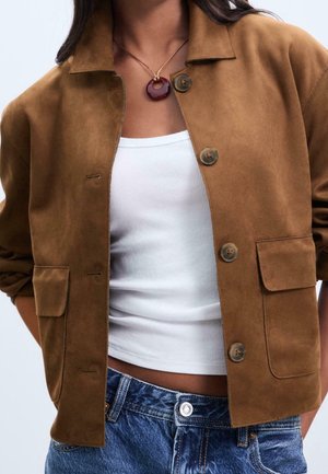 Mujer con chaqueta de ante marrón con botones sobre un top corto blanco de canalé y jeans de mezclilla azules, con un collar redondo con colgante color vino tinto.