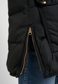 Veste matelassée noire avec fermeture éclair dorée partiellement ouverte sur un jean en denim bleu.