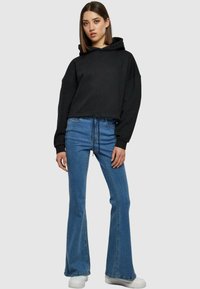 Felpa nera cropped con cappuccio e coulisse, abbinata a jeans svasati blu. L'outfit presenta un design casual e un materiale morbido. Sneakers bianche completano il look.