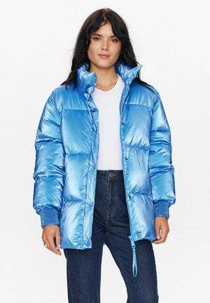 Blauwe pufferjas van glanzende, reflecterende stof, met een hoge kraag, drukknoopsluiting en geribde manchetten. Gedragen over een witte top en jeans.