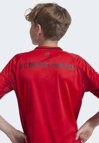 Maillot rouge du FC Bayern München à manches courtes, avec un logo noir et un texte au dos, texture lisse et coupe athlétique.