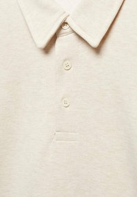 Polo color beige con cuello, tres botones, tejido suave, textura vertical y un discreto detalle de puntada en el bolsillo.