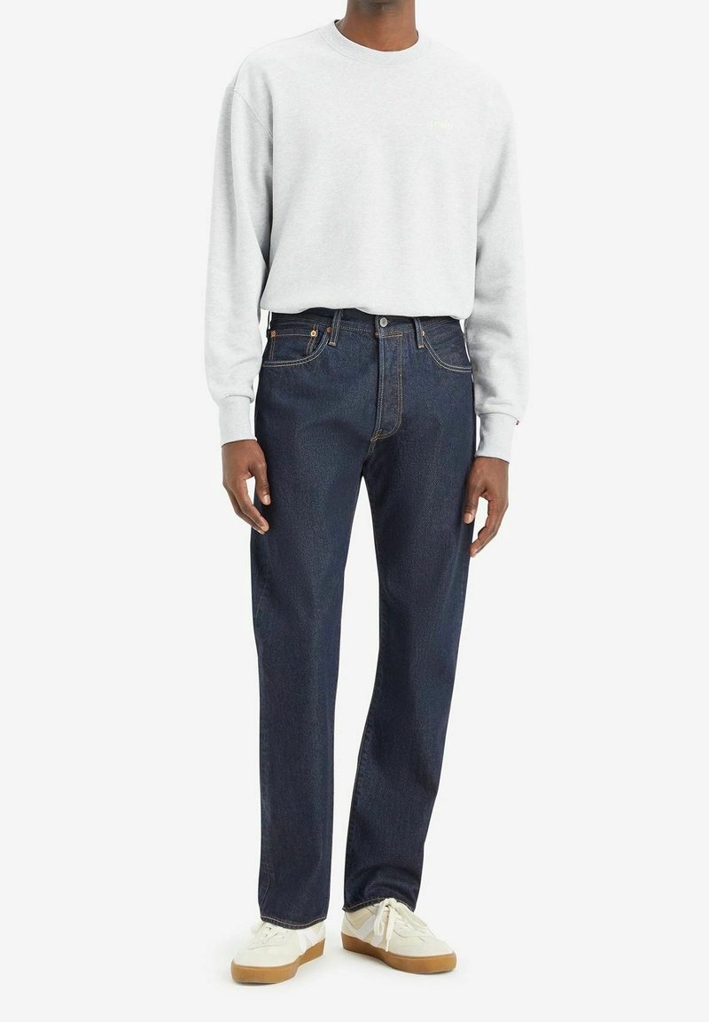 Homme portant un sweat-shirt gris clair, un jean droit bleu foncé et des baskets blanches à semelles gomme, debout devant un fond blanc.