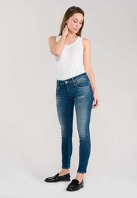 Jeans skinny in denim blu medio con dettagli strappati; abbinati a una canotta bianca e scarpe flat nere. La modella posa con una mano sul collo.