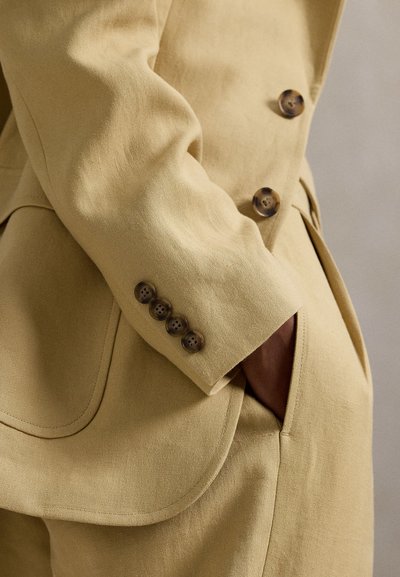 Κίτρινο λινό σακάκι με εφαρμοστή γραμμή, τέσσερα καφέ κουμπιά στη μανσέτα και λεπτομέρεια στην τσέπη. Ομαλή υφή με διακριτική λάμψη.