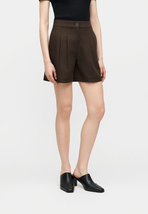 Femme portant un short plissé taille haute brun foncé, un haut noir moulant et des chaussures à talons noires sans lacets, debout devant un fond blanc uni.