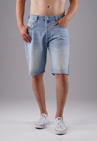 Light blue denim shorts with a straight cut, five pockets, and contrast stitching. Worn with white sneakers, finished with a raw hem.