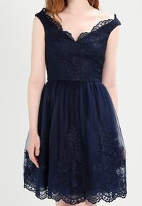 Navy blue dress with a fitted bodice, lace trim, and floral embroidery. Skirt is layered tulle with scalloped lace hem. Sleeveless design.