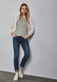 Veste bomber beige clair, t-shirt vert, jeans bleu foncé et baskets blanches avec des accents texturés, se tenant contre un fond gris clair.