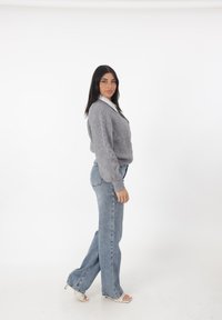 Grau gestrickter Pullover mit V-Ausschnitt, kombiniert mit hellblauen Schlagjeans. Das Outfit beinhaltet ein weißes Hemd mit Kragen darunter.