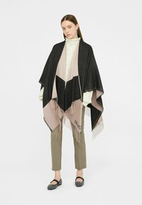 Poncho geometrico nero e rosa con bordi frangiati, realizzato in tessuto morbido. Il modello lo indossa sopra un maglione chiaro e pantaloni grigi, con ballerine.