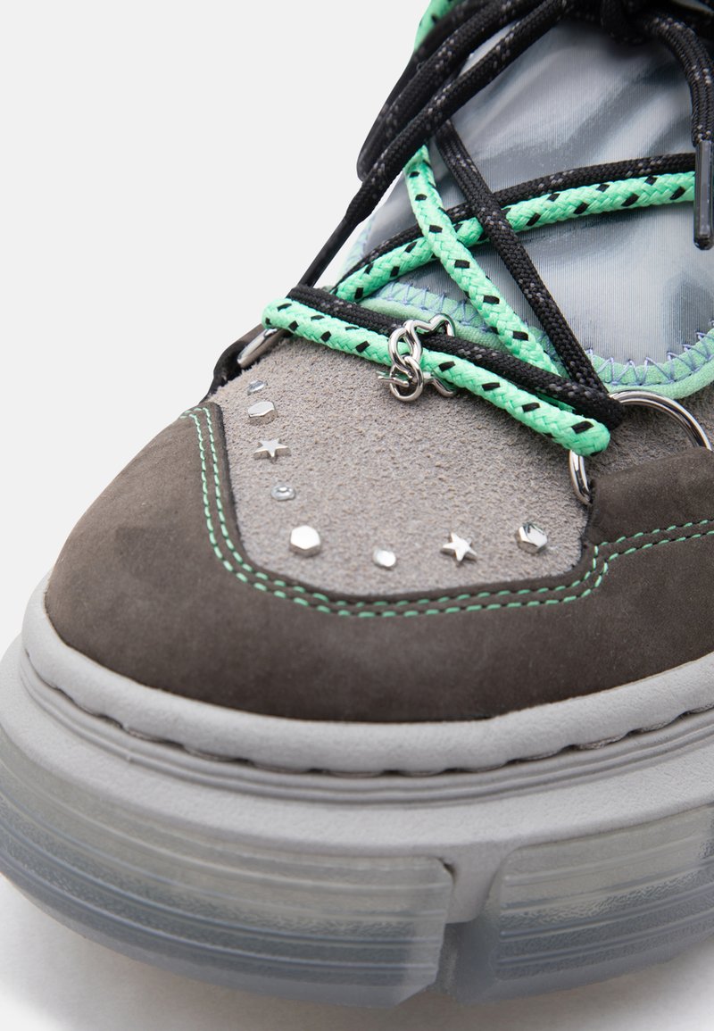 Nahaufnahme eines grauen und mintgrünen Sneakers mit schwarzen und mintfarbenen Schnürsenkeln, metallischen sternförmigen Nieten und dicker durchsichtiger Sohle.