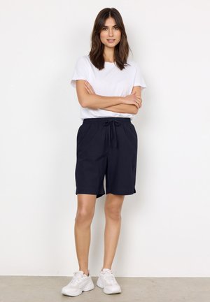 Femme debout, bras croisés, portant un t-shirt blanc, un short bleu marine avec cordon et des baskets blanches sur un fond blanc uni.