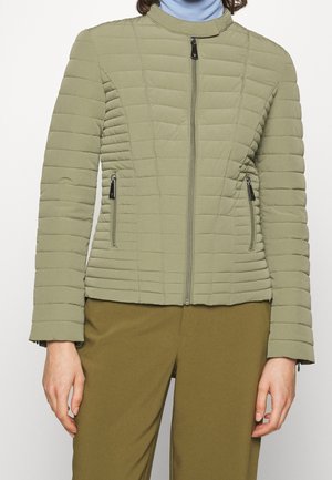 Personne portant une veste matelassée vert clair zippée avec deux poches latérales zippées et un pantalon vert olive.