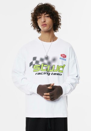 Weißes Langarmshirt mit schwarzen und neongrünen Renngrafiken, ergänzt durch fingerlose braune Strickhandschuhe, vor einem einfarbigen Hintergrund.