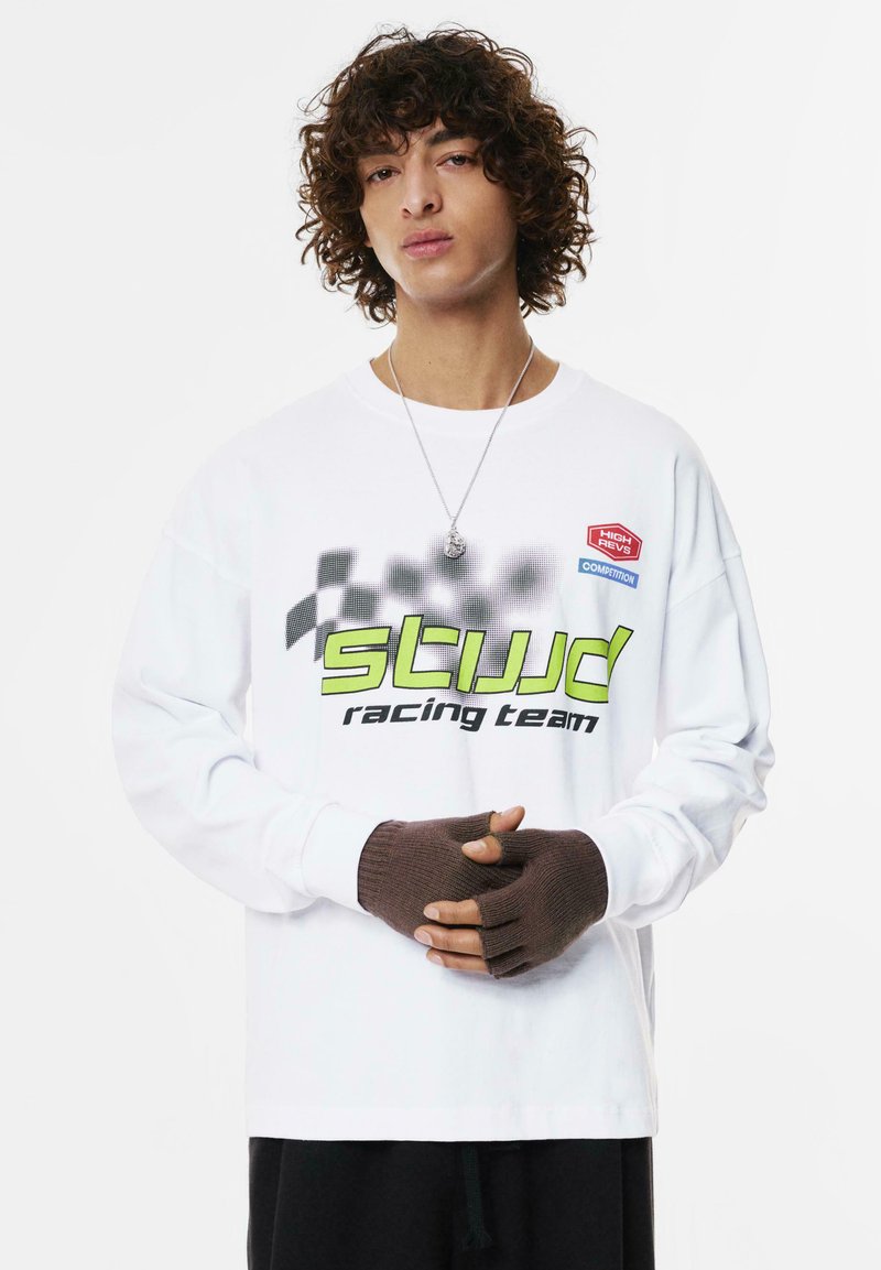 Weißes Langarmshirt mit schwarzen und neongrünen Renngrafiken, ergänzt durch fingerlose braune Strickhandschuhe, vor einem einfarbigen Hintergrund.