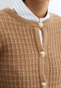 Brązowy dziany kardigan o teksturowanym wzorze, z okrągłym dekoltem i dwoma dekoracyjnymi guzikami z przodu, narzucony na bluzkę w prążki.
