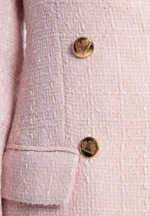 Rosa tweed blazer detail met een textuurstof en een patroon van verhoogde garens, en twee gouden knopen met emblemen.