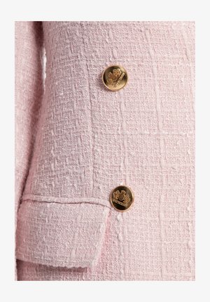 Rosa Tweed-Blazer-Detail mit strukturiertem Stoff und einem Muster aus erhabenen Fäden sowie zwei goldenen Knöpfen mit Emblemen.
