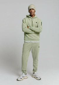 Limoengroene hoodie en joggingset, met een logo op de borst en de linkerarm. G gedragen met een crèmekleurige gebreide beanie en witte sneakers.