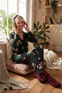 Jonge vrouw in berenprint-pyjama en bordeauxroze sokken zit op een vloerkussen, lacht en draagt een koptelefoon in een gezellige, met planten gevulde kamer.