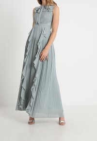 Pitkä, hihaton mekko vaaleanharmaana, jossa on pystysuoria laskoksia ja röyhelökoristeita molemmilla puolilla. Tasaisen sileä kangas ja vyötäisiltä istuva malli.