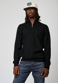 Sweat à zip noir avec manches longues, poignets et ourlet côtelés, porté sur un jean bleu clair, associé à une casquette crème avec des détails graphiques.